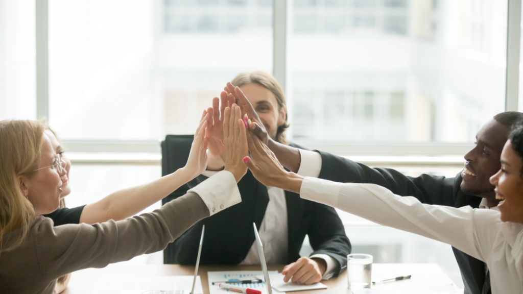 Grupa ludzi uczestniczy w&nbsp;oficjalnym spotkaniu.&nbsp;Spotkanie przebiega w&nbsp;radosnej atmosferze.&nbsp;Ludzie zbijają sobie piątki nad stołem.