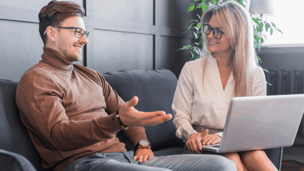Uśmiechnięty mężczyzna w&nbsp;brązowym golfie rozmawia z kobietą siedzącą obok,&nbsp;która trzyma laptop na kolanach i&nbsp;również się uśmiecha;&nbsp;oboje siedzą na kanapie w&nbsp;jasnym,&nbsp;nowoczesnym wnętrzu,&nbsp;co sugeruje spotkanie coachingowe lub konsultacyjne.
