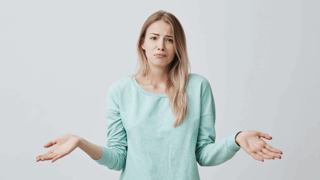 Na zdjęciu widoczna jest młoda kobieta o jasnych,&nbsp;długich włosach,&nbsp;ubrana w&nbsp;miętową bluzę z długim rękawem,&nbsp;stojąca na tle jasnej,&nbsp;jednolitej ściany.&nbsp;Jej postura oraz wyraz dłoni,&nbsp;lekko uniesionych i&nbsp;rozłożonych na boki,&nbsp;mogą symbolizować niepewność,&nbsp;konsternację lub zastanowienie—uczucia często towarzyszące osobom doświadczającym syndromu oszusta.