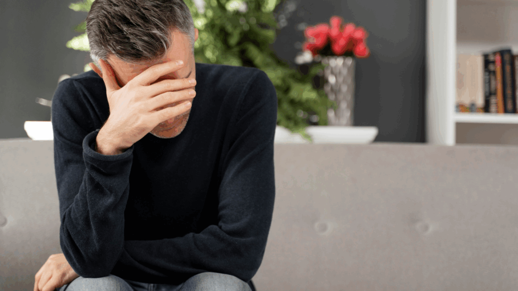 Wypalenie zawodowe – przytłoczony i&nbsp;zmęczony mężczyzna siedzi na kanapie,&nbsp;zasłaniając twarz dłonią w&nbsp;geście przytłoczenia.