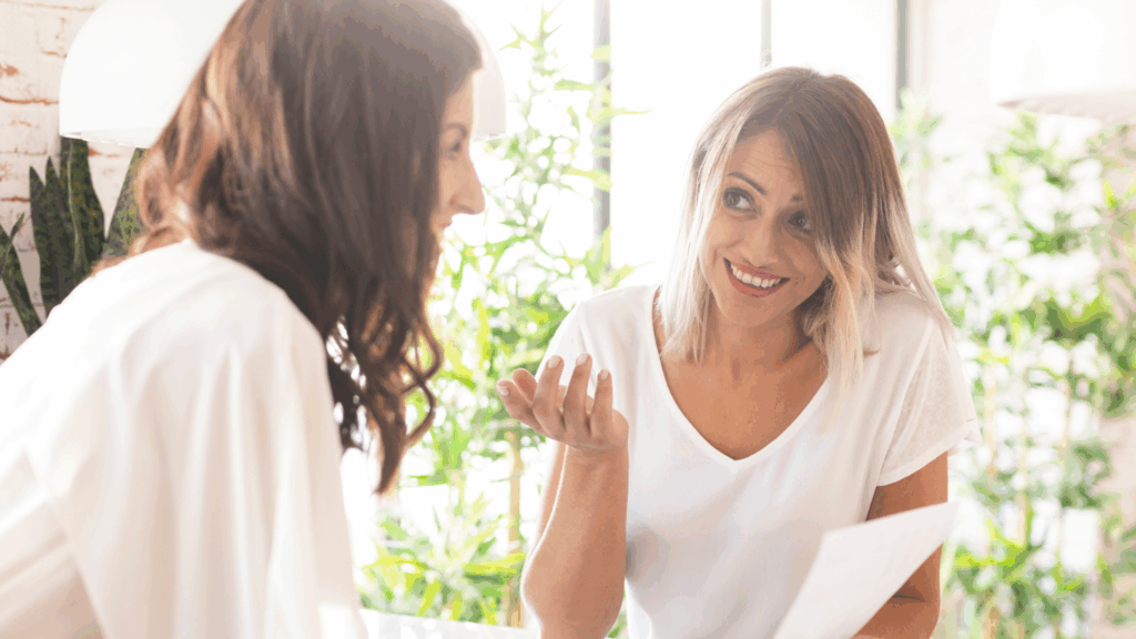 Dwie kobiety rozmawiające w&nbsp;jasnym,&nbsp;roślinnym wnętrzu;&nbsp;jedna uśmiecha się i&nbsp;gestykuluje,&nbsp;trzymając kartkę papieru.
