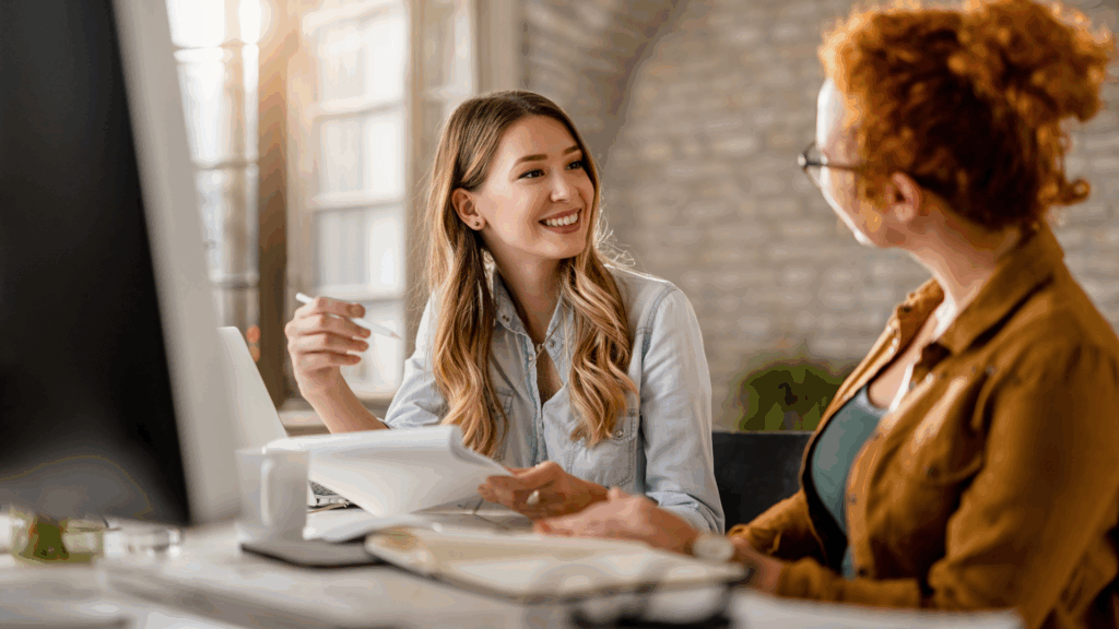 Dwie kobiety współpracujące w&nbsp;kreatywnym biurze;&nbsp;jedna pokazuje notatki drugiej podczas rozmowy.