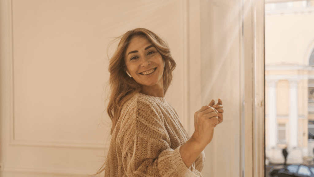 Uśmiechnięta kobieta emanująca pewnością siebie,&nbsp;stojąca w&nbsp;jasnym pomieszczeniu przy oknie.